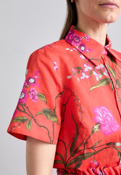 Red floral shirt with short sleeves, featuring vibrant pink and purple flowers and green leaves. Collared design with button accents.