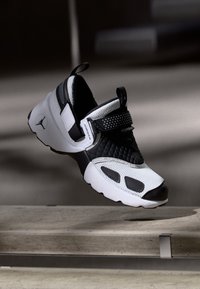 Αθλητικό παπούτσι σε ασπρόμαυρο χρώμα με υφή στο πάνω μέρος, στοιχεία από δίχτυ, επένδυση στον αστράγαλο και λουράκι με Velcro. Διαθέτει εμφανές λογότυπο στο πλάι.