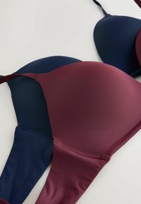 Twee bh's zijn op elkaar gestapeld, een bordeauxrood en een marineblauw. Beide hebben een gladde stof met vormgegeven cups en verstelbare bandjes.