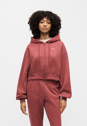 Kobieta z kręconymi włosami ubrana w rdzawą bluzę Nike na zamek oraz dopasowane spodnie dresowe na jednolitym białym tle.