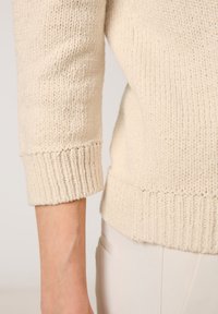 Beige gestrickter Pullover mit strukturiertem Stoff und gerippten Bündchen. Der Saum ist ebenfalls gerippt, was einen lässigen Schnitt betont.
