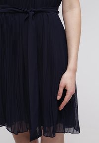 Merenkatu pleidattu mekko, jossa on sileä, kevyt rakenne, itsetaitettava vyö ja virtaavat hihat, esittelee kerroksellista kangasta.