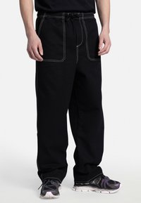 Pantaloni della tuta neri con cuciture bianche, dotati di una vita con cordino e ampie tasche anteriori. Indossati con sneakers nere e viola con texture.
