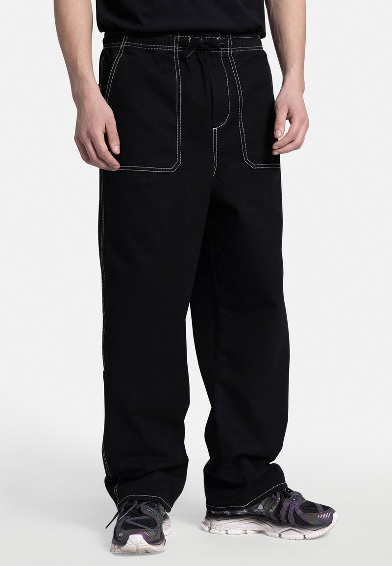Pantaloni della tuta neri con cuciture bianche, dotati di una vita con cordino e ampie tasche anteriori. Indossati con sneakers nere e viola con texture.