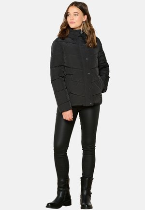Doudoune noire avec des coutures horizontales, un col montant et des fermetures à pression sur le devant. Portée avec un pantalon noir ajusté et des bottines.