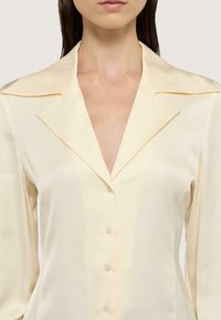 Cremefarvet skjorte med knaplukning, skarp krave, glat stof og seks knapper foran.