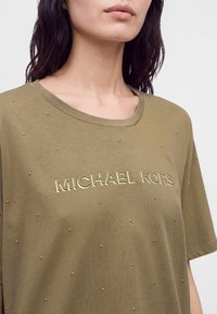 Άτομο που φοράει ελιάς πράσινο μπλουζάκι Michael Kors με μικρά χρυσά τρουκς διάσπαρτα στο ύφασμα και στρογγυλή λαιμόκοψη.
