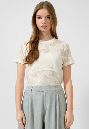 Młoda kobieta z długimi jasnobrązowymi włosami, ubrana w przejrzystą białą koronkową bluzkę z kwiatowym motywem z krótkim rękawem oraz wysokowysokie szare spodnie w prążki.