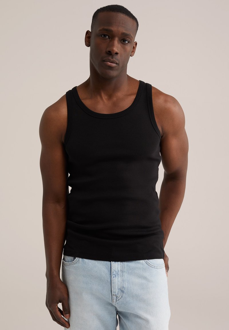 Mężczyzna w czarnej, bezrękawnikowej koszulce i jasnoniebieskich dżinsach stoi na jednolitym tle.