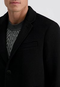 Zwarte wollen blazer van een wolmix met gekartelde revers, enkele knoopsluiting en details van externe zakken. Grijs gebreid vest zichtbaar eronder.