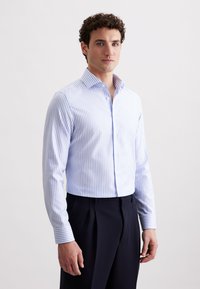 Camicia a maniche lunghe a righe blu e bianche, con design a bottoni, in cotone, colletto aperto, silhouette aderente, abbinata a pantaloni scuri.