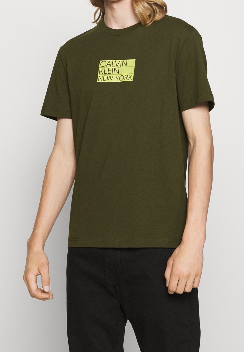Tmavě olivově zelené bavlněné tričko s krátkými rukávy, na kterém je uprostřed hrudi žluté grafické logo "CALVIN KLEIN NEW YORK".