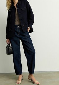 Mørkeblå tekstureret jakke med frontlommer, beige tanktop, mørkeblå løst siddende jeans og nude højhælede sandaler, mens du holder en lille sort taske.