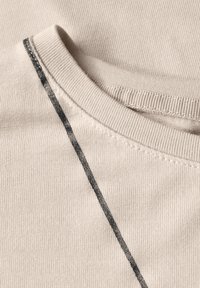 Beige bavlněná látka s vroubkovanou strukturou. Obsahuje černý akcent poblíž švu. Detailní záběr na kulatý výstřih a ramenní části.