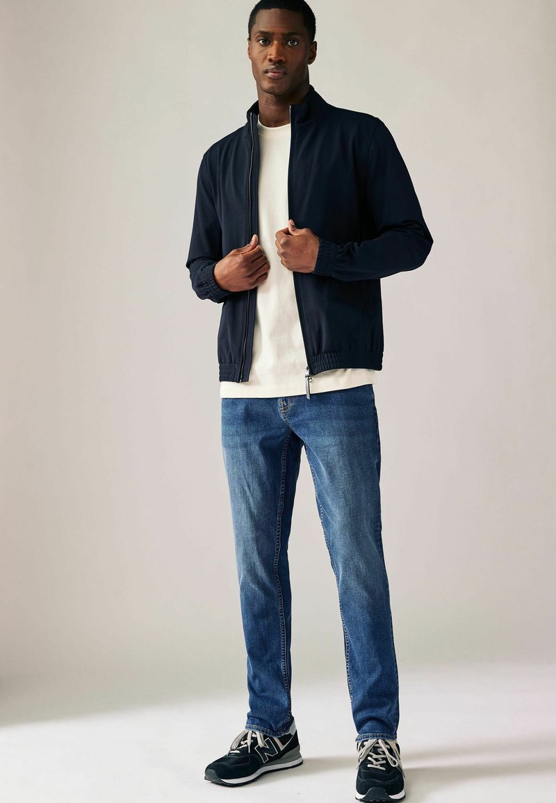 Marine-zipjack met geribde manchetten, gecombineerd met een witte t-shirt en blauwe jeans. Zwarte sportieve sneakers met witte zolen maken de look compleet.