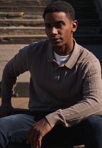 Světle hnědý žebrovaný polo tričko s límečkem, kombinované s tmavě modrými džíny. Model sedí na schodech za přirozeného světla a s texturovaným pozadím.