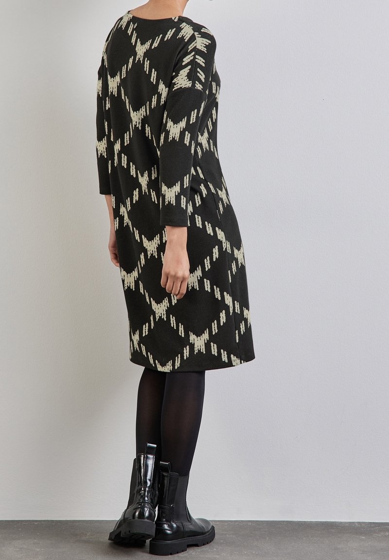 Robe noire en tricot avec un motif abstrait crème, manches trois quarts et coupe décontractée, associée à des bottes noires montantes.