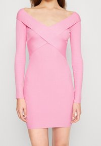 Donna che indossa un vestito rosa aderente a maniche lunghe con scollatura a incrocio scoperta sulle spalle, in piedi contro uno sfondo neutro.