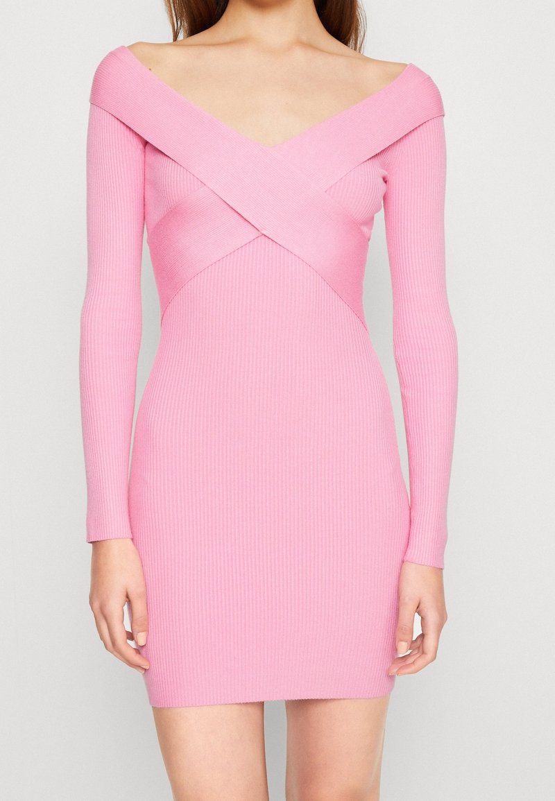 Donna che indossa un vestito rosa aderente a maniche lunghe con scollatura a incrocio scoperta sulle spalle, in piedi contro uno sfondo neutro.