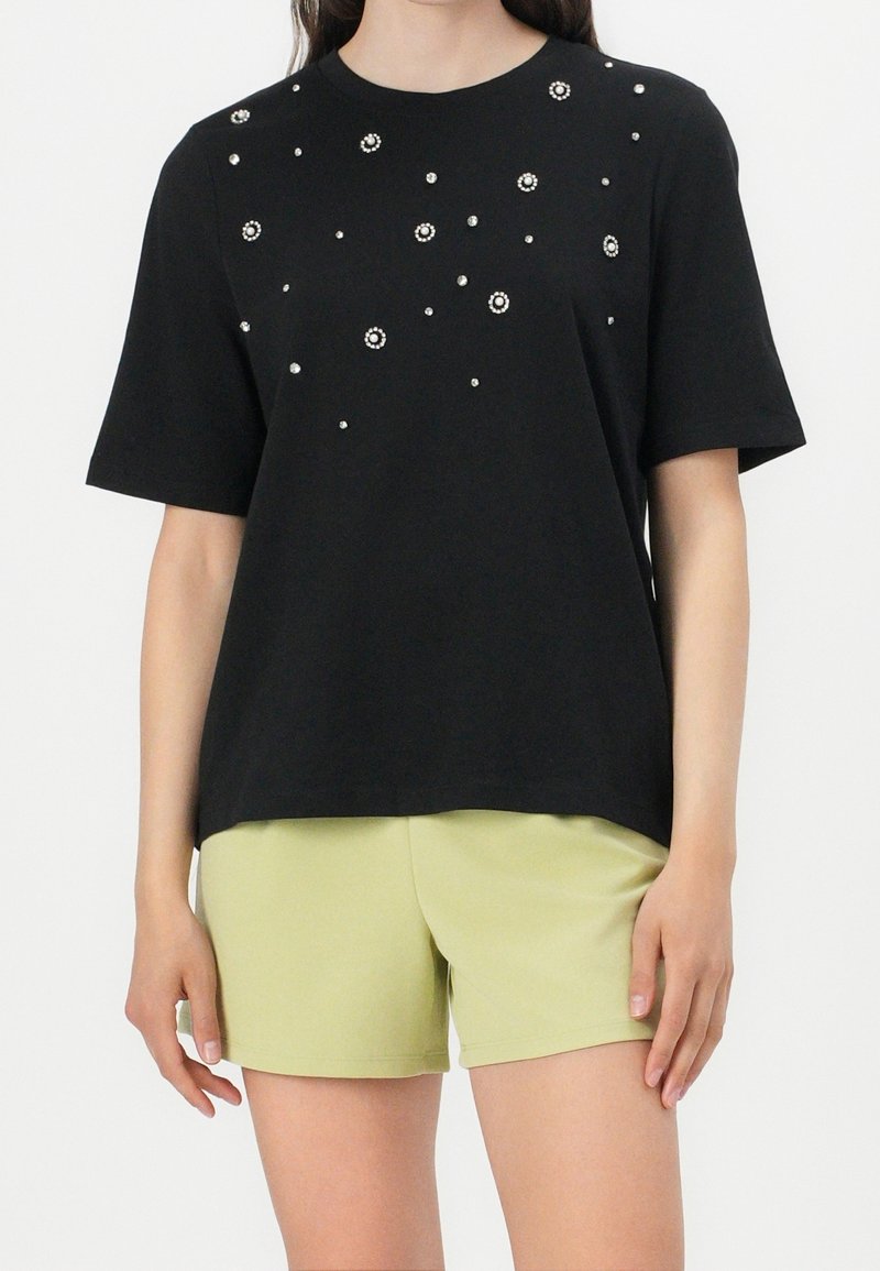 T-shirt noir à manches courtes avec des embellissements texturés, comportant des clous en argent dispersés et une coupe ample, associé à un short vert clair.