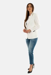 Blazer blanco con botones negros sobre una camisa blanca, combinado con jeans ajustados de color azul claro y tacones altos negros. Tejido suave y diseño estructurado.