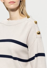 Heller beiger Pullover mit marineblauen Streifen, geripptem Halsauschnitt und dekorativen goldenen Knöpfen an der Schulter. Strickstruktur, lockere Passform.