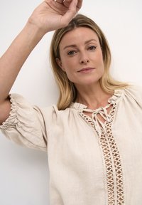 Beige len anyagú felső V-kivágással, bonyolult csipkés díszítéssel és puffos, gumis mandzsettás ujjaival. A nyakban lévő masnik teljesítik ki a dizájnt.