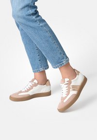 Personne portant un jean bleu clair et des baskets beige-blanc avec des lacets blancs, en plein mouvement sur un fond blanc uni.