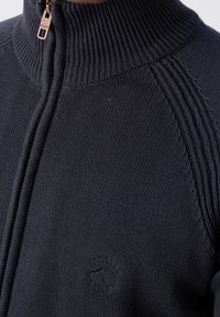 Gros plan d'un pull en tricot noir zippé avec col côtelé et logo en relief sur la poitrine sous l'épaule gauche.