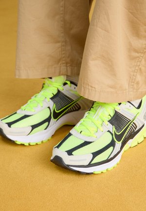 Baskets Nike vert néon et noir portées avec un pantalon beige sur une surface jaune.