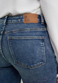 Jeans de mezclilla azul con una cintura alta, que presentan una etiqueta de cuero en la parte trasera con "BLUE RIDGE" en relieve y un diseño clásico de bolsillos.