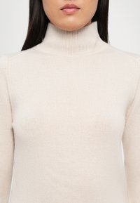 Lys beige høyhalset genser laget av mykt strikket stoff, med en figursydd silhuett og ribbestruktur ved kragen og ermene.