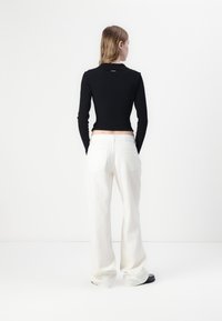 Maglietta nera a coste con maniche lunghe e design aderente, abbinata a pantaloni bianchi oversize e scarpe nere. La vista posteriore mostra una vita alta e due tasche posteriori.