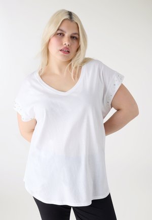 Kobieta z jasnoblond włosami nosi biały krótki top z perełkami i koralikami na rękawach oraz czarne spodnie, na białym tle.