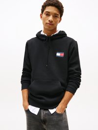 Junger Mann in einem schwarzen Tommy Jeans Hoodie über einem weißen Hemd, mit den Händen in den Taschen, steht vor einem schlichten hellen Hintergrund.