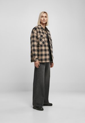 Chemise en flanelle écossaise beige et noire, avec deux poches poitrine, associée à un jean noir à large jambe et des chaussures noires. Texture lisse.