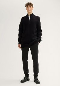 Schwarzer Strick-Zip-Pullover mit hohem Kragen, strukturiertem Muster, kombiniert mit schlanken schwarzen Hosen und schwarzen Schnürschuhen.