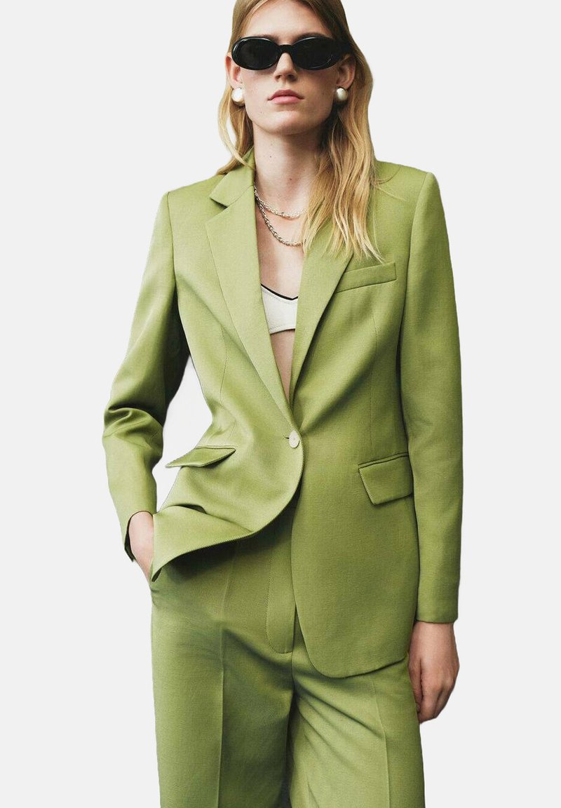 Groene blazer en bijpassende broek van textuurstof. De blazer heeft een enkele knoop, notch-panden en een borstzak.