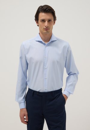 Man wearing a light blue and white striped button-up shirt with long sleeves and navy blue trousers, standing with one hand in pocket.