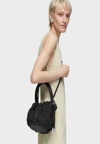 Bolso tejido negro con forma redondeada, que presenta dos asas de cuero y detalles en acero inoxidable, colocado sobre un fondo neutro.