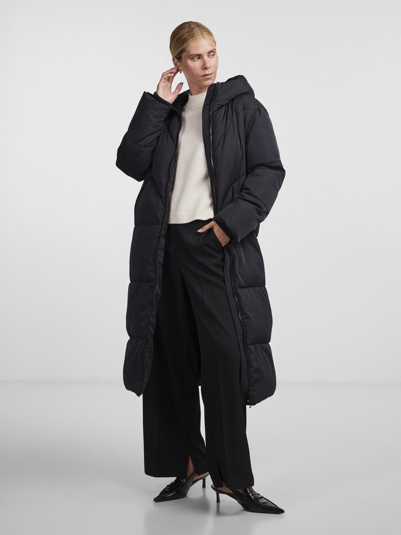 Black padded long coat with a hood, featuring a front zipper, side pockets, and a glossy finish. Paired with a cream sweater and black trousers.