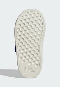 Podešev bílého sandálu Adidas s šestiúhelníkovým vzorem a logem Adidas blízko paty, umístěná na světle šedém pozadí.