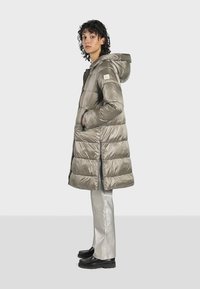 Cappotto piumino grigio chiaro con cappuccio, design trapuntato, tasche laterali e dettagli riflettenti nella zip, abbinato a pantaloni metallici argento.