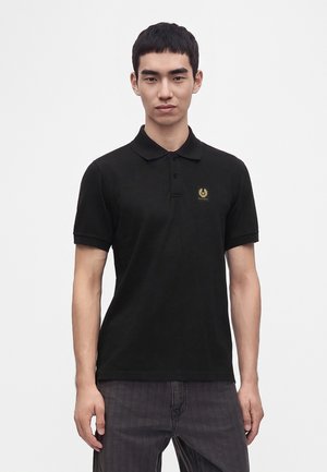 Junger Mann trägt ein schwarzes, kurzärmeliges Polohemd mit einem kleinen goldenen Logo auf der linken Brust und dunkelgraue Hose, steht vor weißem Hintergrund.