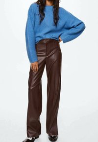 Person, der einen blauen gerippten Pullover trägt, kombiniert mit hochgeschnittenen, weit geschnittenen braunen Lederhosen, steht mit einer Hand auf der Hüfte.