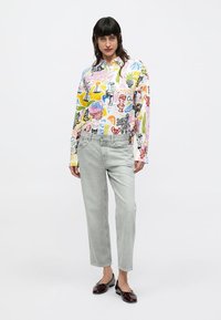 Chemise à manches longues blanche avec des motifs graphiques colorés ; associée à un jean gris clair à jambes droites et des chaussures en cuir verni foncé.