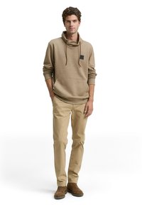 Beige hoodie met een col en een enkele borstzak, gecombineerd met lichtbruine broeken en bruine enkellaarzen, tegen een witte achtergrond.