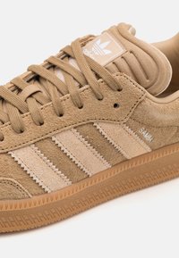 Tan mockasiner i semsket skinn med lysebeige tre striper, gummisåle og polstret krage. Har en merkeetikett på tungen og teksturerte detaljer.