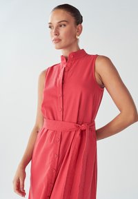 Korálové bezrukávové šaty s vysokým golierom, predným zapínaním na gombíky a opasok v páse. Vyrobené z ľahkej látky s hladkou textúrou.