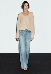 Jasno beżowy sweter dzianinowy z dekoltem w kształcie litery V i ribbed dolnym wykończeniem, zestawiony z luźnymi, spranymi niebieskimi dżinsami. Modelka stoi na ciemnej powierzchni na tle białej ściany.